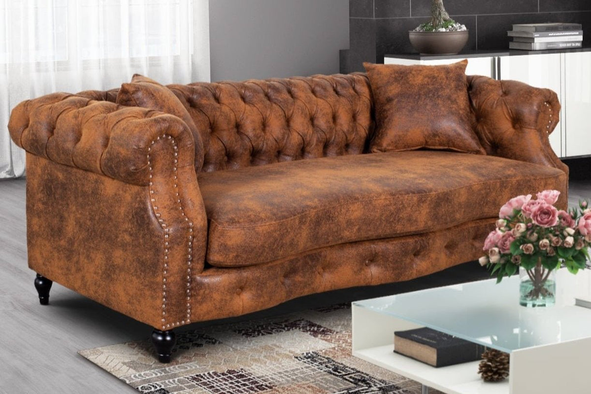 A brown faux leather suede Richmond 3 seater Chesterfield sofa with deep buttoning and decorative studding on the armrests. The sofa is set on a hardwood frame with elegant black legs and comes with two matching accent cushions.