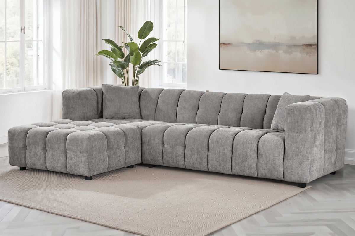 An L-shaped tufted sofa with left hand facing chaise end section, upholstered in grey chenille fabric, styled in a bright, modern living room