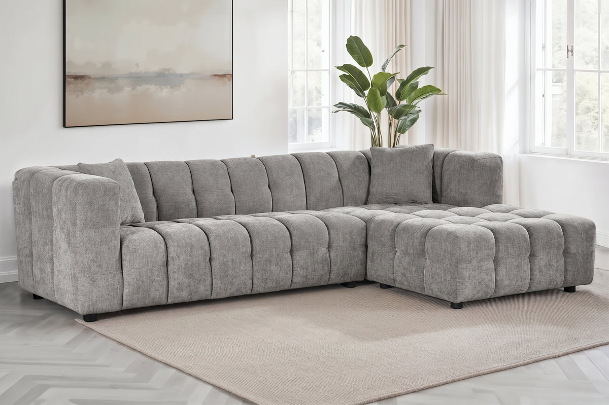 An L-shaped tufted sofa with right hand facing chaise end section, upholstered in grey chenille fabric, styled in a bright, modern living room